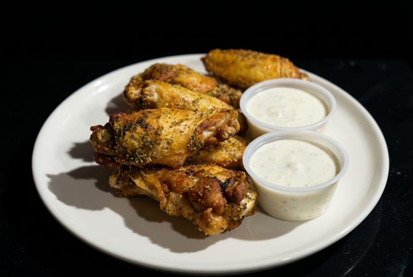 A white plate holds several seasoned chicken wings and two small cups of ranch dressing.