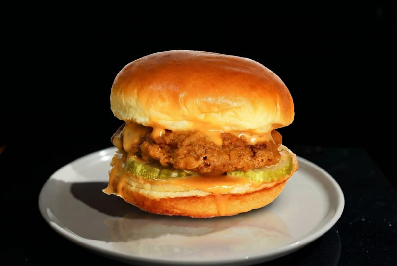 Crispy fried chicken sandwich with pickles and orange sauce on a brioche bun, on a white plate.