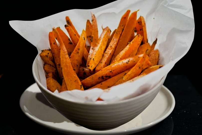 A white bowl filled with golden-orange sweet potato fries, lined with white parchment paper.