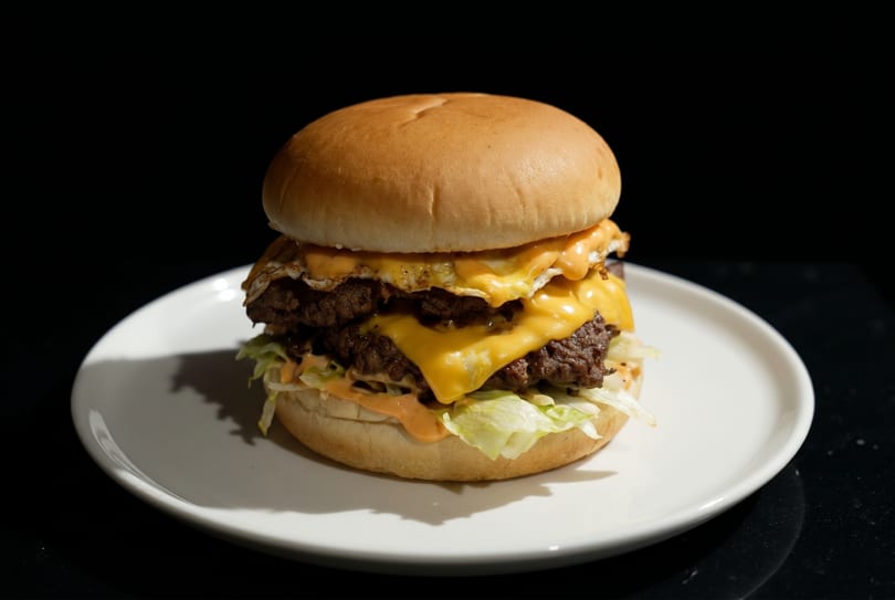 A double cheeseburger with fried eggs, lettuce, and sauce on a white plate against a dark background.