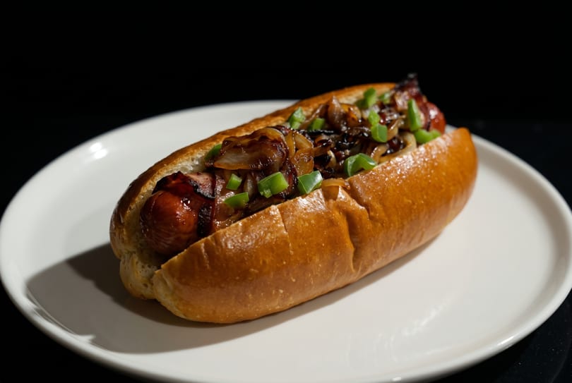 Grilled hot dog in a bun topped with caramelized onions and chopped green onions on a white plate.