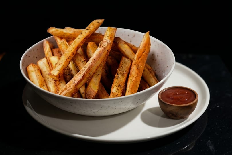A bowl of seasoned french fries and a small wooden cup of ketchup on a white plate.