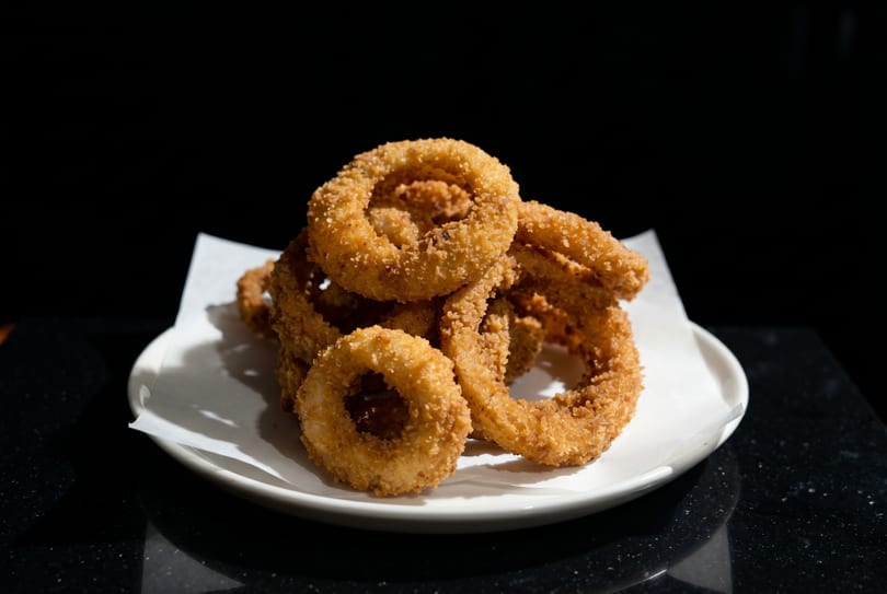 A white plate piled high with golden-brown, crispy onion rings on a dark surface.