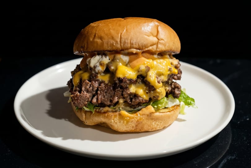 A double cheeseburger with melted cheese, lettuce, and sauce on a white plate against a dark background.